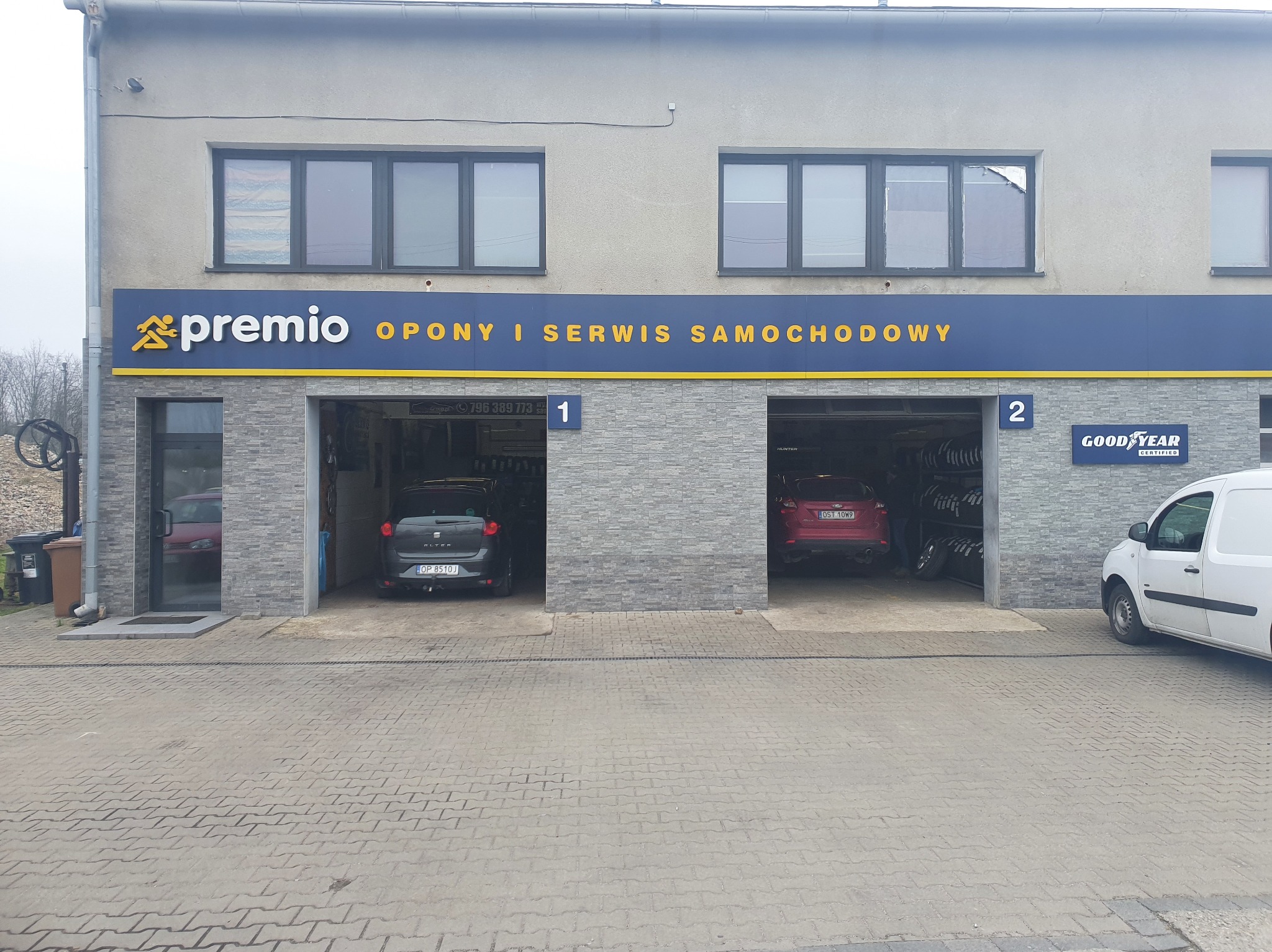 Budynek serwisu Premio z dwoma stanowiskami, oponami i zaparkowanymi autami. Widoczny szyld z numerem telefonu i logo Goodyear. Szara kostka brukowa na podjeździe.