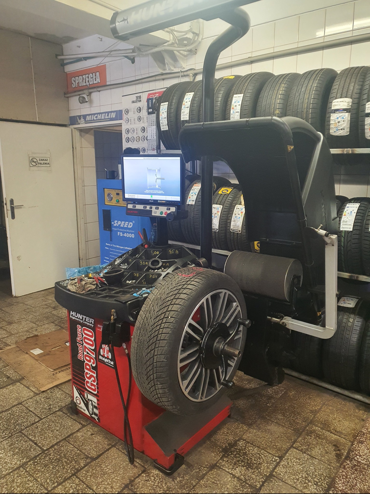 Wyważarka Hunter GSP9700 z oponą na stanowisku, monitor z symulacją wyważania, w tle rząd opon na półkach i tablica Michelin w warsztacie samochodowym.