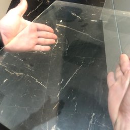 Cleaning Time - Dłonie sprawdzają czystość szklanego blatu na ciemnym, marmurkowym tle po umyciu. Widoczny metalowy pojemnik w tle.