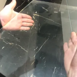 Dłonie sprawdzają czystość szklanego blatu na ciemnym, marmurkowym tle po umyciu. Widoczny metalowy pojemnik w tle.