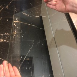 Cleaning Time - Dwie dłonie trzymające szklaną taflę nad błyszczącą, czarną podłogą z marmurowym wzorem, obok szarej szafki kuchennej.