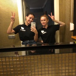 Cleaning Time - Dwie uśmiechnięte kobiety w czarnych koszulkach polo z logo firmy sprzątającej pozują do selfie w łazience z błyszczącymi złotymi kafelkami, obie pokazują znak 'V'.