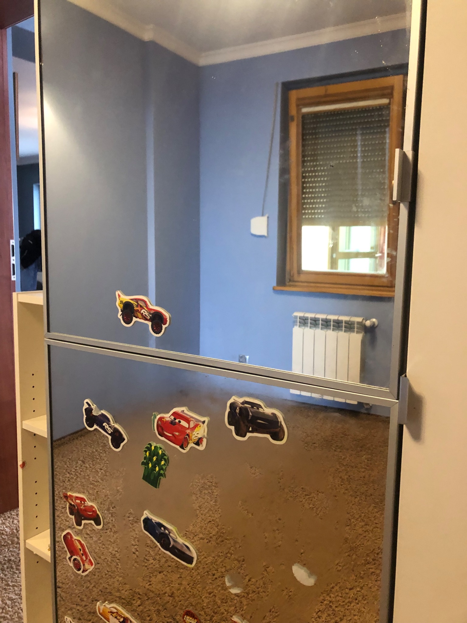 Powierzchnia lustra oklejona naklejkami z motywem samochodów, odbijająca pokój z niebieskimi ścianami, oknem z roletą i białym kaloryferem.