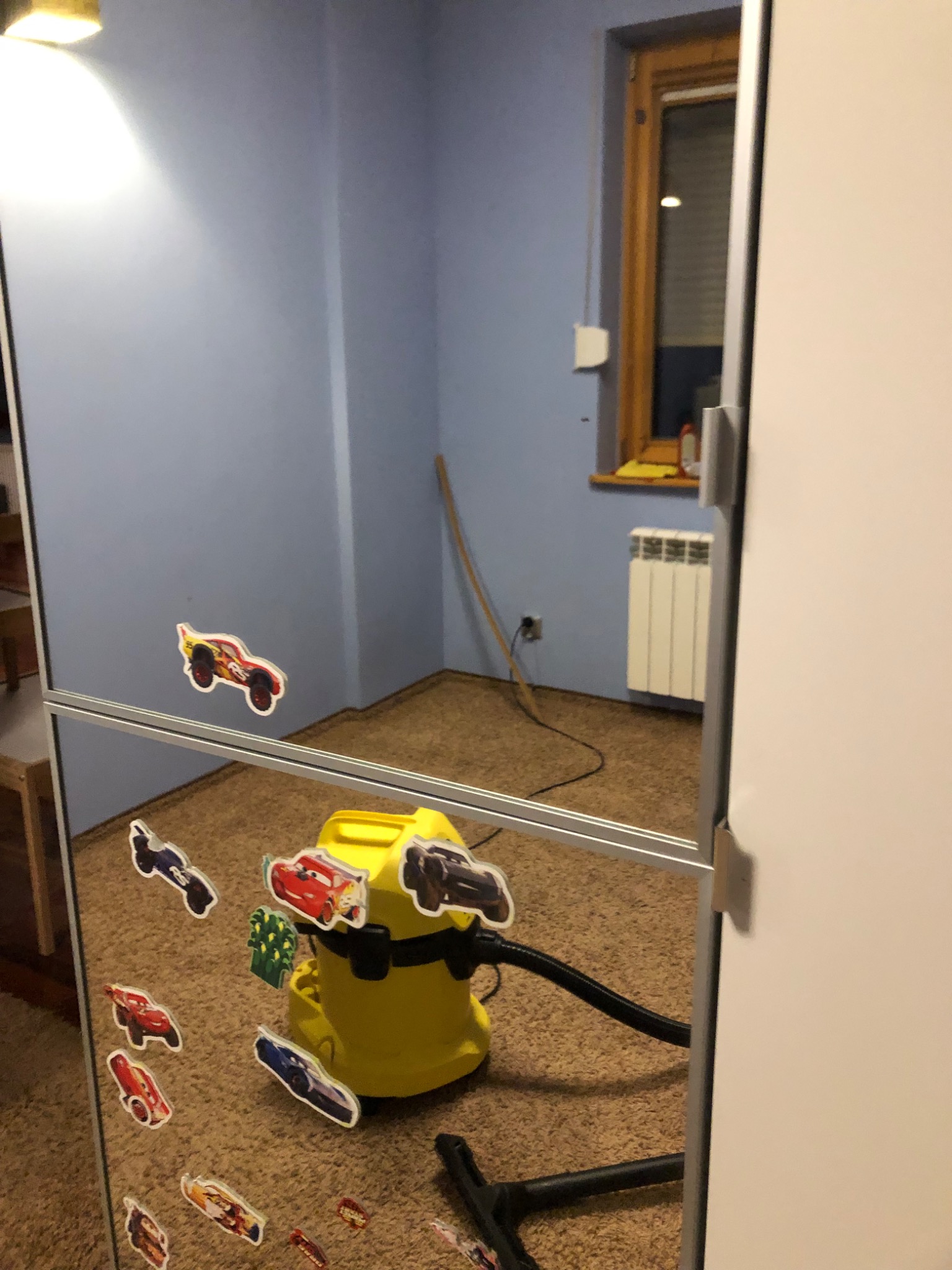 Żółty odkurzacz piorący, widoczny w odbiciu lustra, ozdobionego naklejkami z motywami samochodów, na tle pokoju z brązowym dywanem i niebieskimi ścianami.