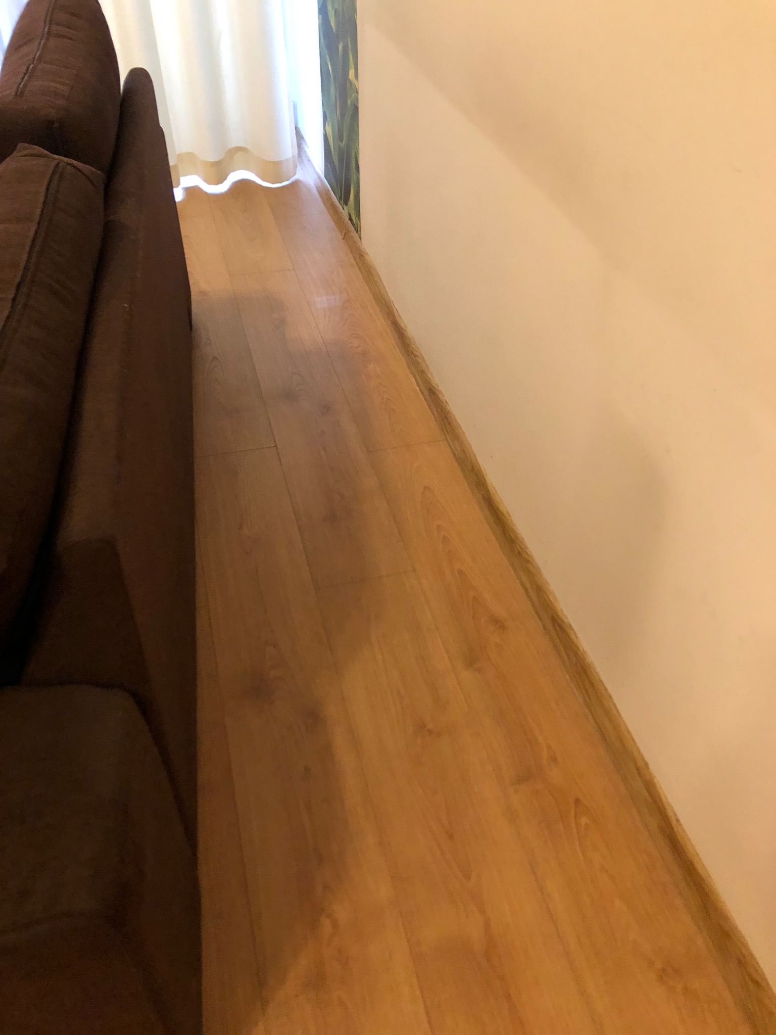 Fragment brązowej kanapy, drewniana podłoga w odcieniach brązu i beżowa ściana z listwą przypodłogową, widoczne zasłony w tle.