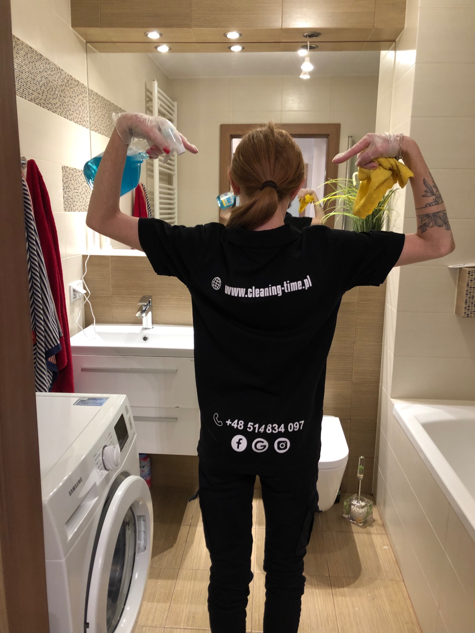 Osoba w czarnej koszulce z logo firmy sprzątającej, stojąca tyłem w łazience, w rękawiczkach, trzymająca w jednej ręce spryskiwacz z niebieskim płynem, a w drugiej żółtą ściereczkę, wskazując...