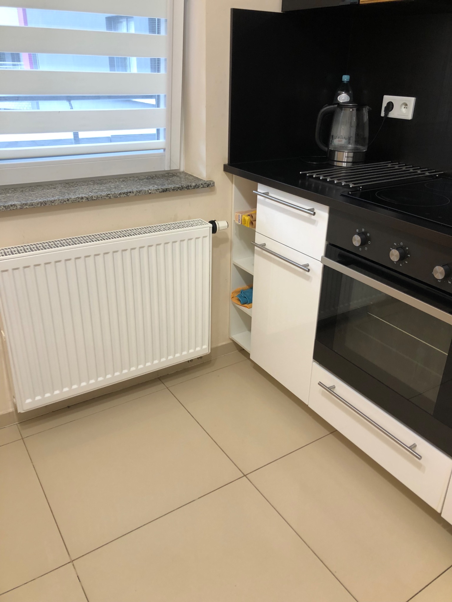 Fragment kuchni z białymi szafkami, piekarnikiem z czarnym frontem, grzejnikiem pod oknem z żaluzjami poziomymi, czajnikiem elektrycznym na blacie.