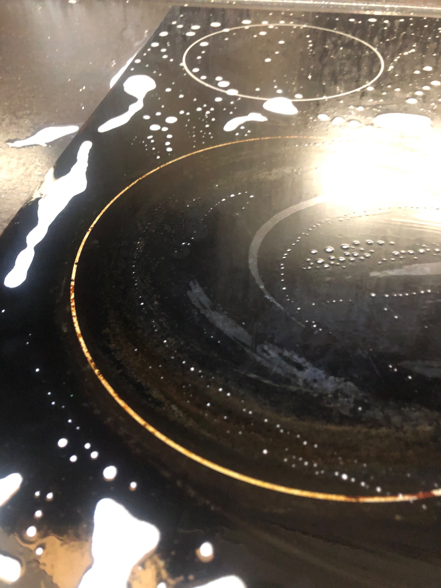 Czyszczenie płyty indukcyjnej: biała piana detergentu na czarnej powierzchni z widocznymi okręgami pól grzewczych, częściowo wytarta, z resztkami zabrudzeń wokół obwodu.