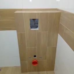 Stelaż podtynkowy WC obudowany płytkami imitującymi drewno, z widocznymi przyłączami i otworem rewizyjnym, na tle białych płytek ściennych.