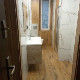 Wykończona łazienka z drewnopodobnymi płytkami na podłodze i ścianach, białą umywalką nablatową i kabiną prysznicową za szybą, widziana zza otwartych drzwi.