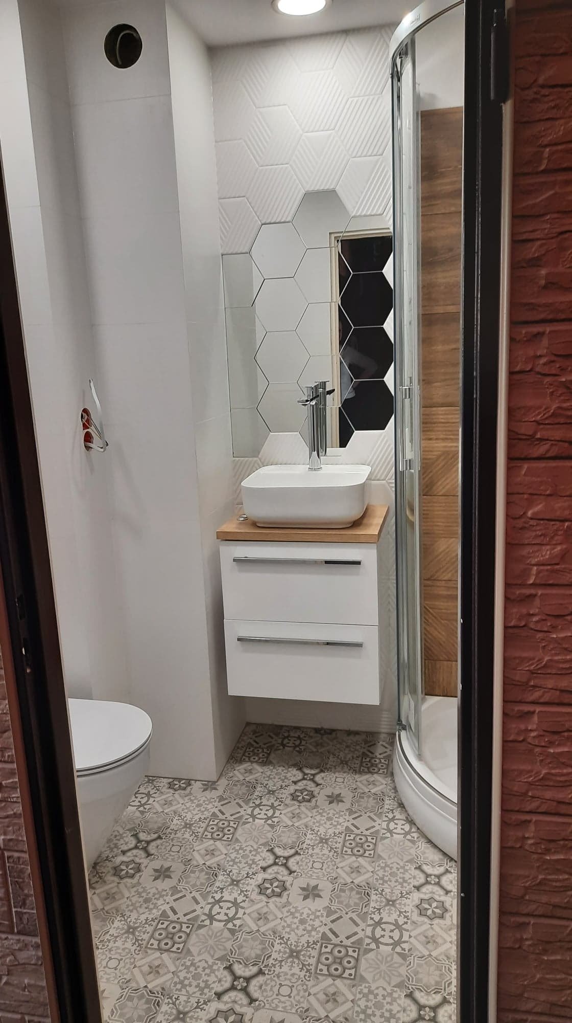 Nowoczesna łazienka po remoncie: białe płytki heksagonalne z lustrzanymi elementami, biała szafka z umywalką na blacie z drewna, kabina prysznicowa z drewnianą wstawką, dekoracyjne płytki podłogowe...