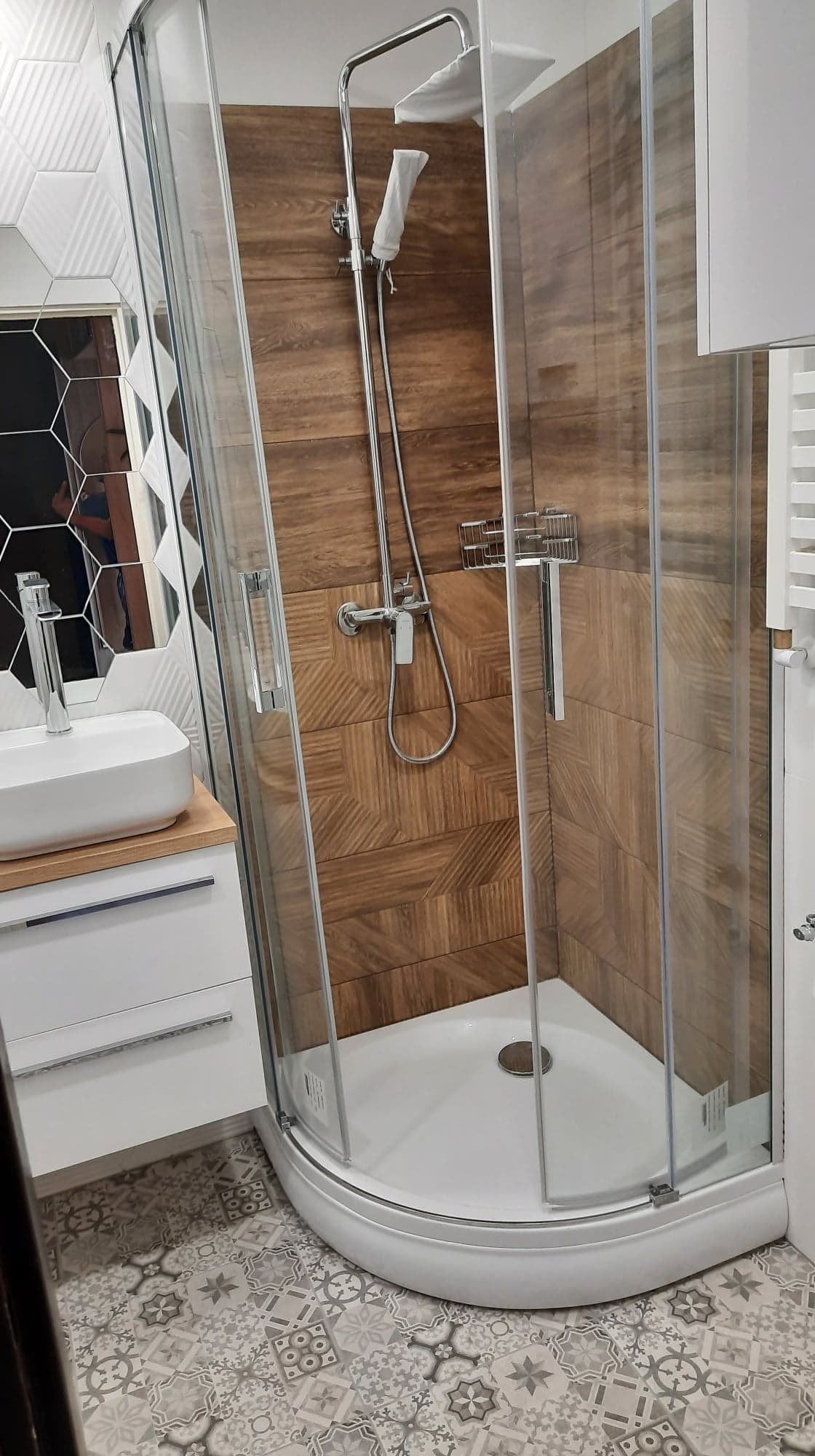 Nowoczesna kabina prysznicowa narożna z transparentnymi szybami, wykończona drewnopodobnymi płytkami ułożonymi we wzór jodełki, z chromowaną armaturą i białą brodziką, uzupełniona o umywalkę...