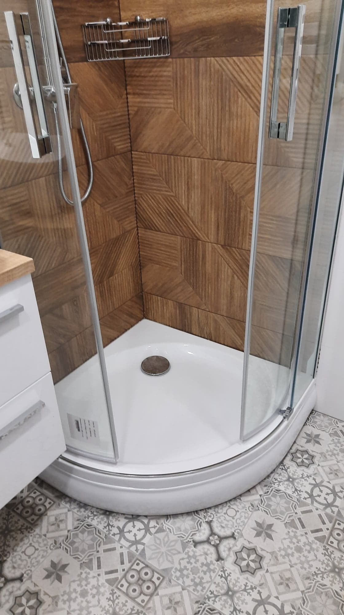 Nowoczesna kabina prysznicowa narożna z drewnopodobnymi płytkami ułożonymi w geometryczny wzór na ścianach i wzorzystymi szarymi płytkami na podłodze, widoczna umywalka z szafką po lewej stronie.