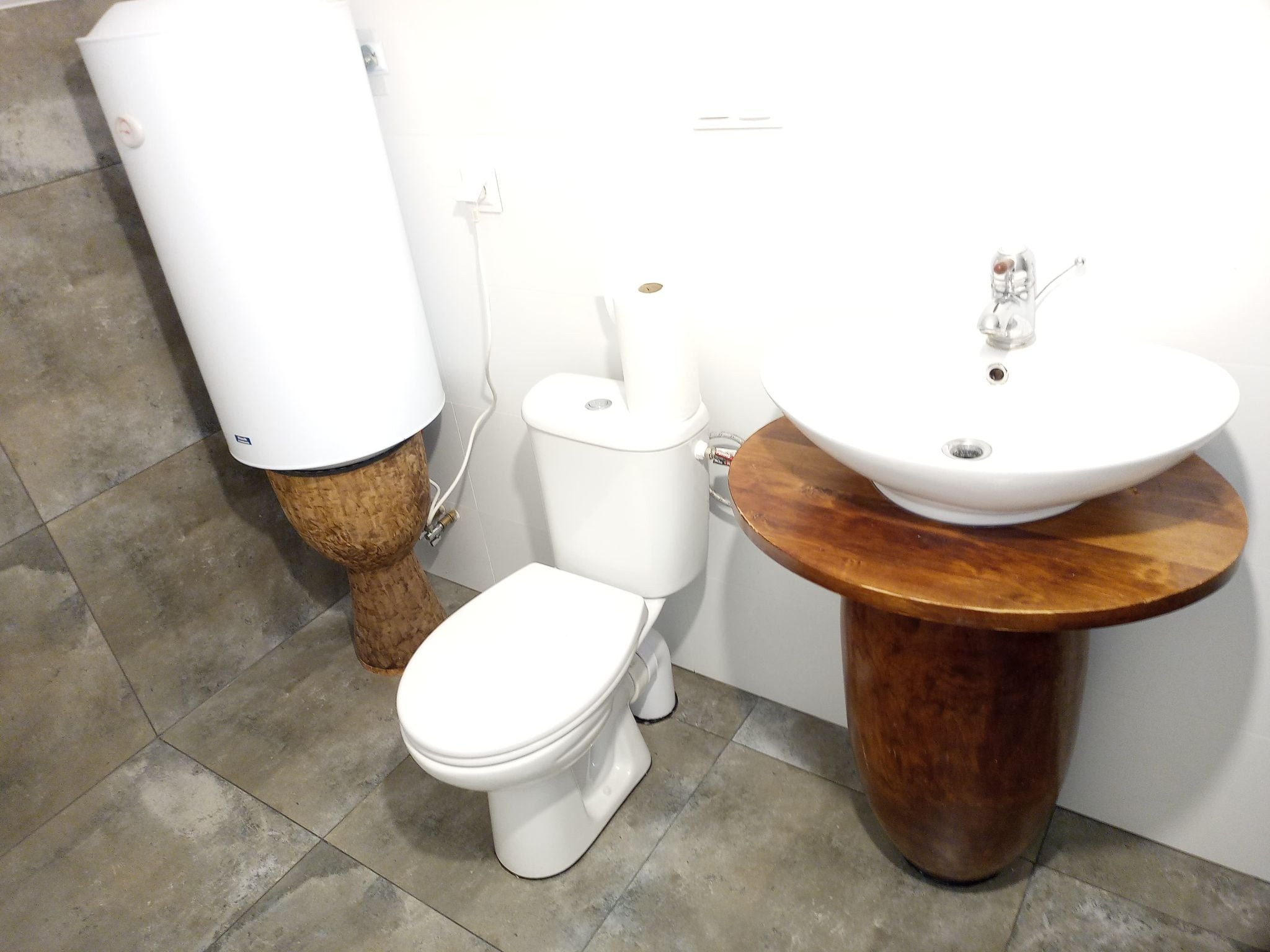 Biała toaleta i umywalka na drewnianym postumencie w łazience wyłożonej szarymi płytkami. Bojler elektryczny na plecionym cokole.