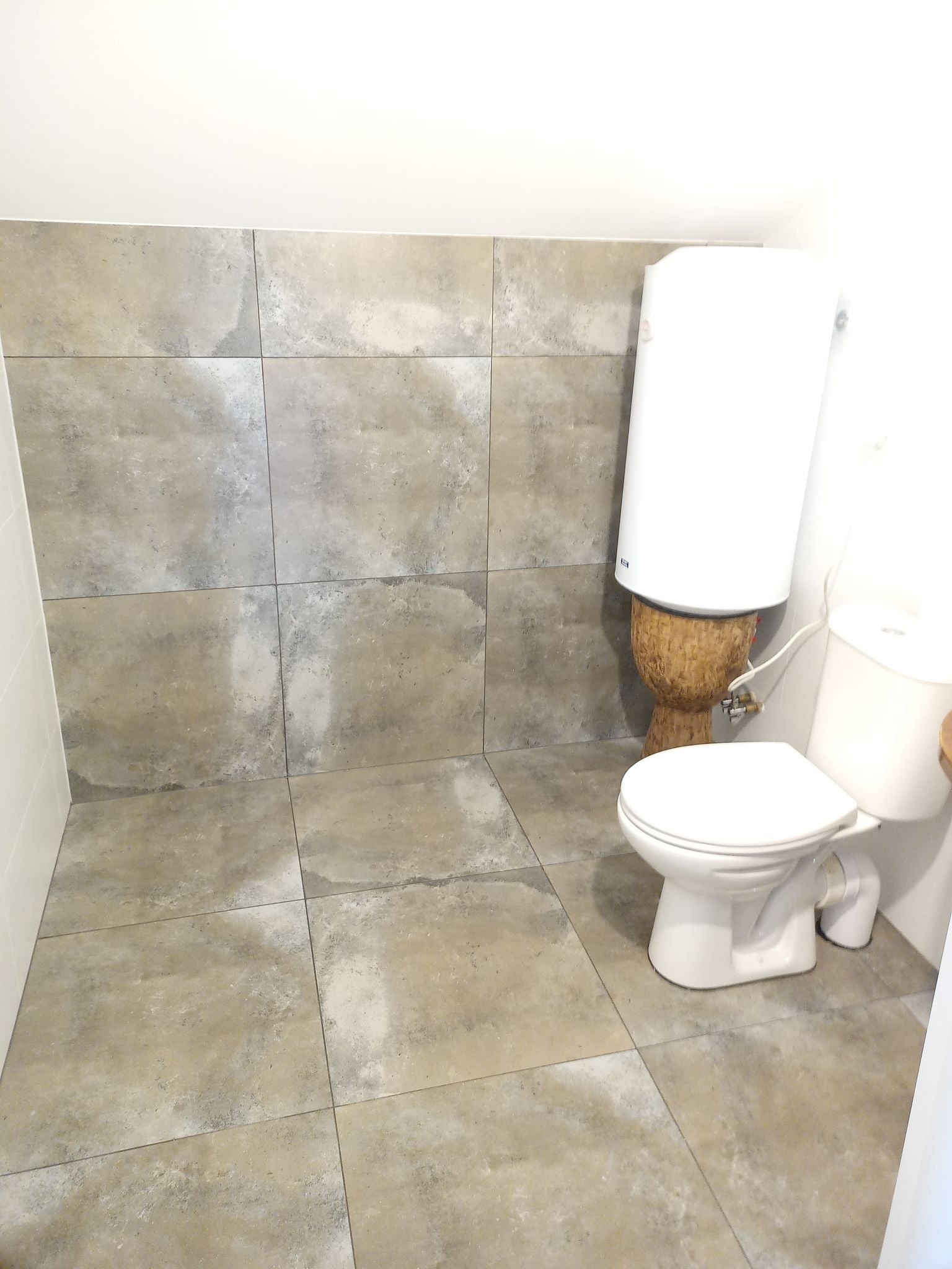 Płytki podłogowe i ścienne w łazience w odcieniach szarości, biała toaleta i bojler zamontowany na drewnianej podstawie w kształcie bębna.