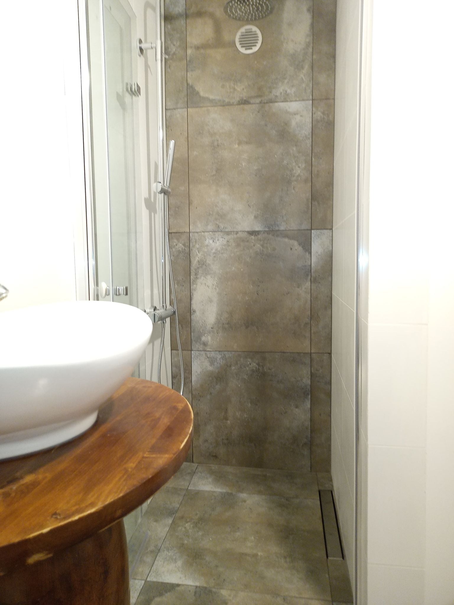 Nowoczesna łazienka z kabiną prysznicową wyłożoną płytkami imitującymi kamień, okrągłą umywalką nablatową na drewnianym blacie i odpływem liniowym w podłodze.