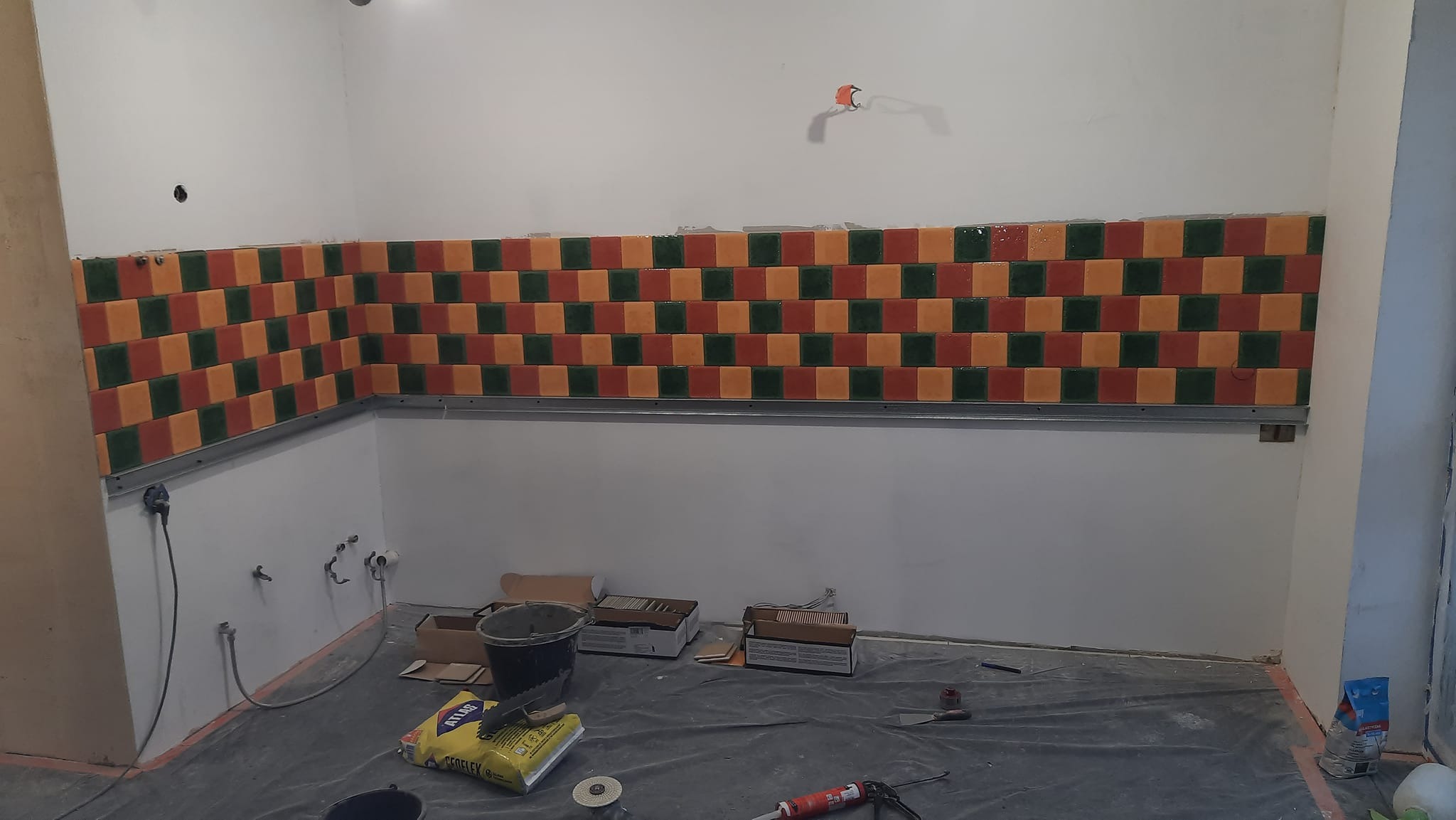 Świeżo ułożone, kolorowe, kwadratowe płytki w odcieniach zieleni, pomarańczy i czerwieni na ścianie w trakcie remontu kuchni lub łazienki, widoczne narzędzia i materiały budowlane na podłodze...