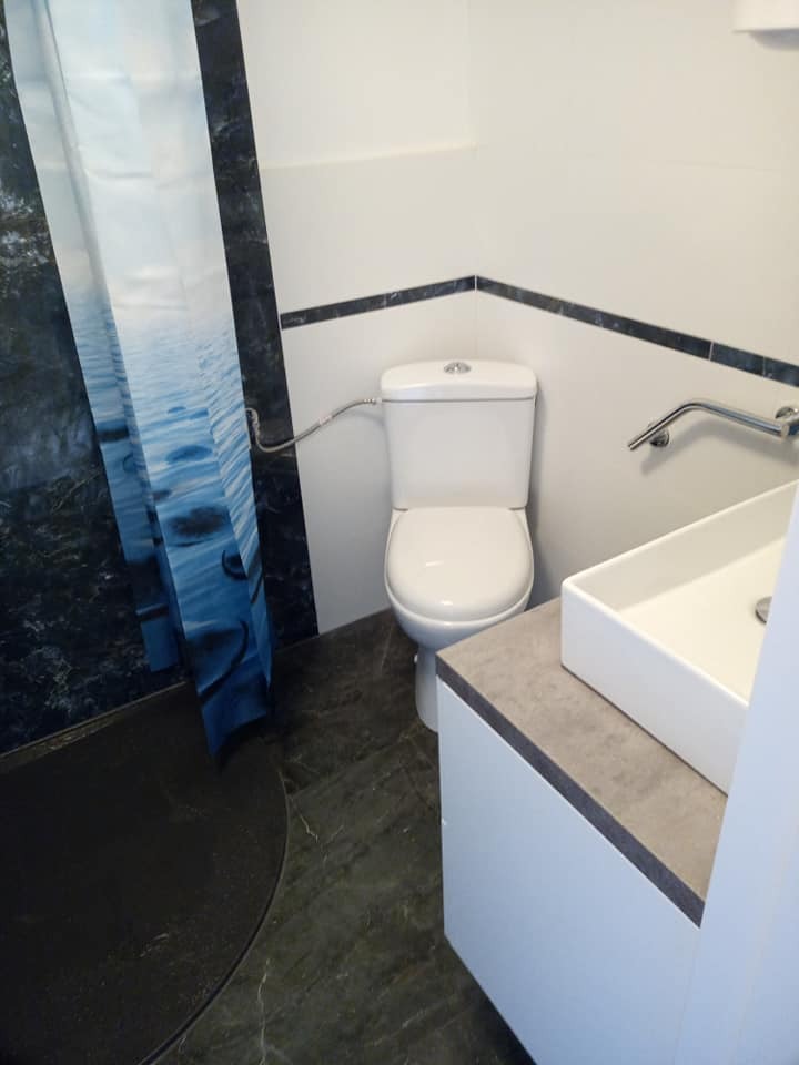 Nowoczesna łazienka z kabiną prysznicową z zasłoną z motywem wody, białą toaletą i umywalką na szafce z blatem w kolorze betonu, wykończona ciemną listwą przy suficie.