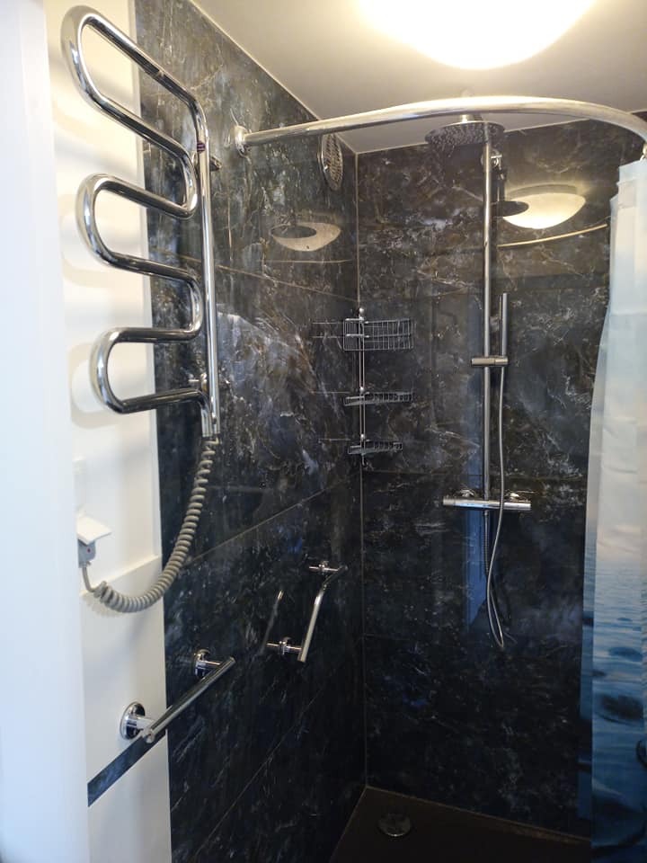 Wykończona kabina prysznicowa z ciemnymi marmurkowymi płytkami, wyposażona w chromowany grzejnik drabinkowy, półki na kosmetyki, baterię i deszczownicę, z widocznym fragmentem zasłony prysznicowej...