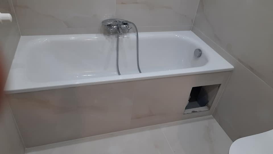 Biała wanna obudowana płytkami w łazience, widoczny fragment instalacji przez otwór rewizyjny.