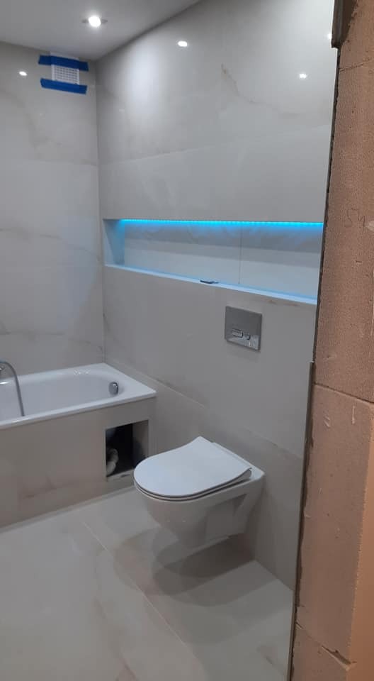 Nowoczesna łazienka z białymi płytkami imitującymi marmur, podświetlaną wnęką nad wanną niebieskim światłem LED, wiszącą toaletą i fragmentem niezabezpieczonego rogu ściany.