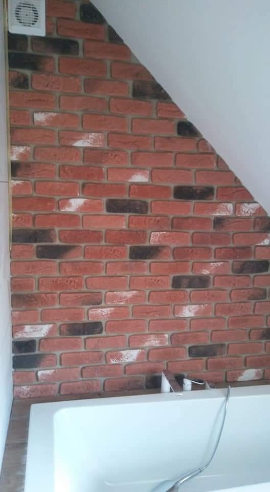 Ściana w łazience wykończona dekoracyjną cegłą w odcieniach czerwieni i brązu, częściowo pod skosem dachu, z widoczną białą wanną i elementem wentylacji.