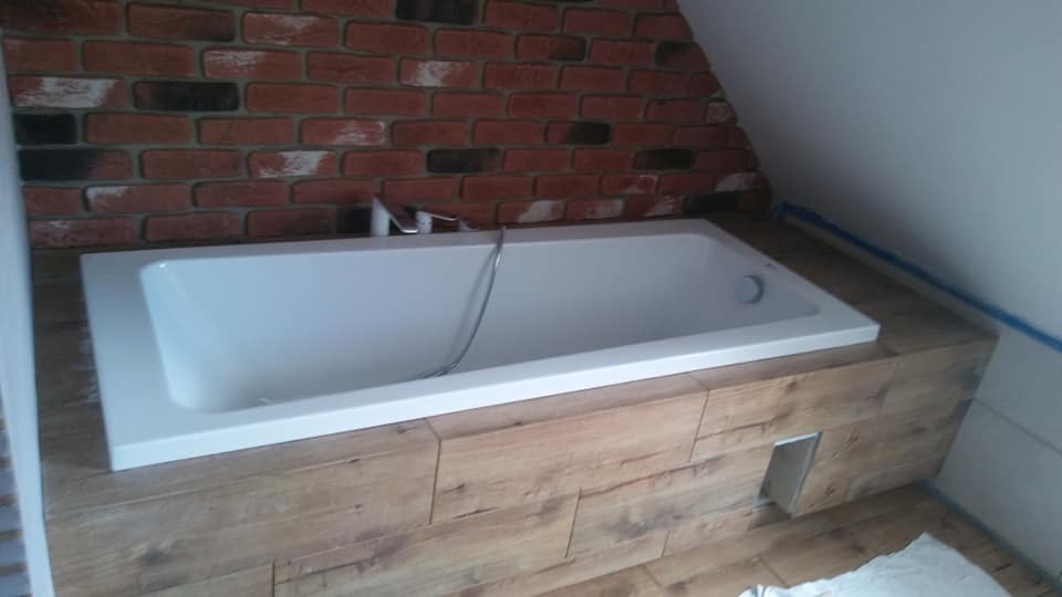 Prostokątna wanna w obudowie z drewnopodobnych paneli, z baterią i słuchawką prysznicową na tle ściany z cegły dekoracyjnej w łazience na poddaszu.