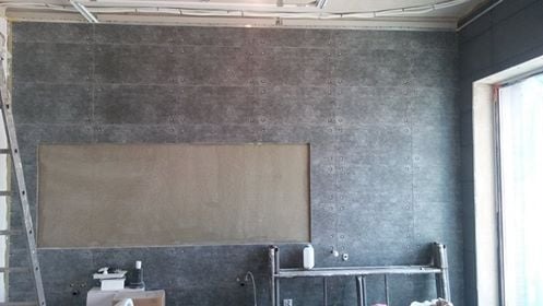 Ściana wyłożona szarymi płytkami z dekoracyjnymi nitami, z prostokątnym otworem przygotowanym do dalszych prac wykończeniowych, widoczna drabina i elementy instalacji pod sufitem.