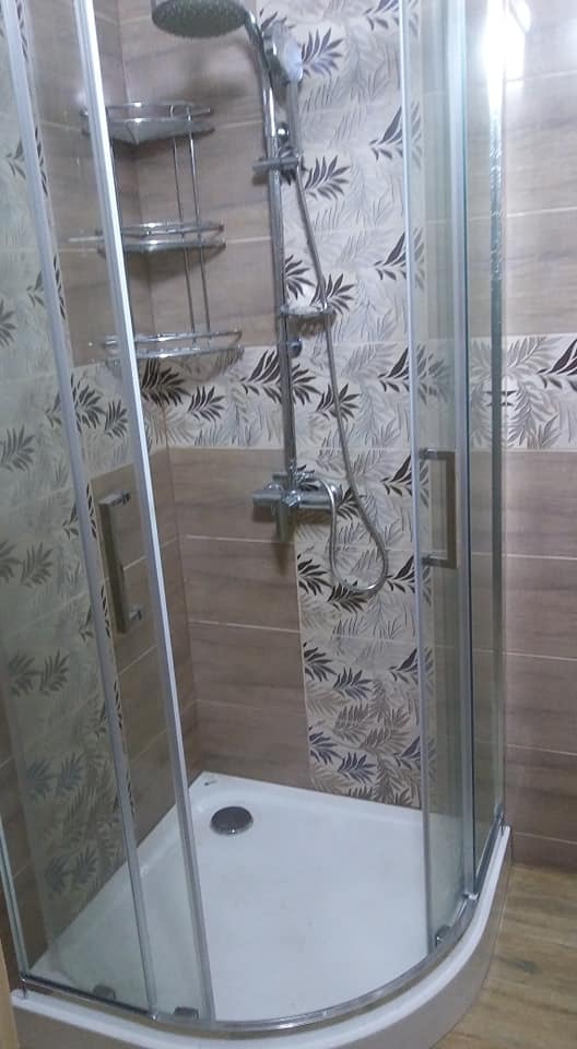 Zmontowana kabina prysznicowa z chromowaną armaturą i półkami narożnymi, wykończona płytkami w odcieniach beżu i szarości z motywem liści.