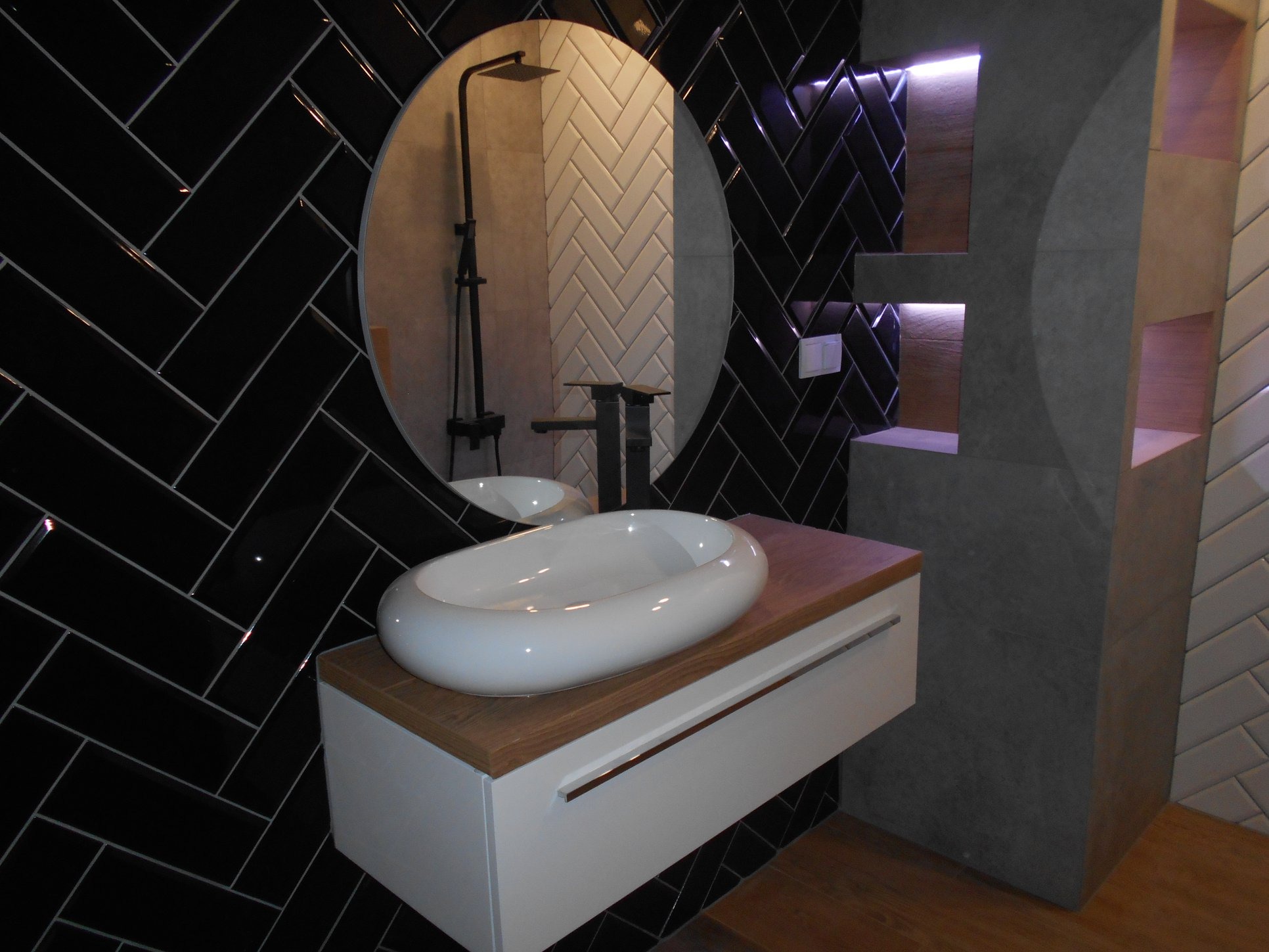 Nowoczesna łazienka z czarnymi płytkami ułożonymi w jodełkę, owalną białą umywalką na drewnianym blacie i okrągłym lustrem odbijającym kabinę prysznicową z czarną armaturą.