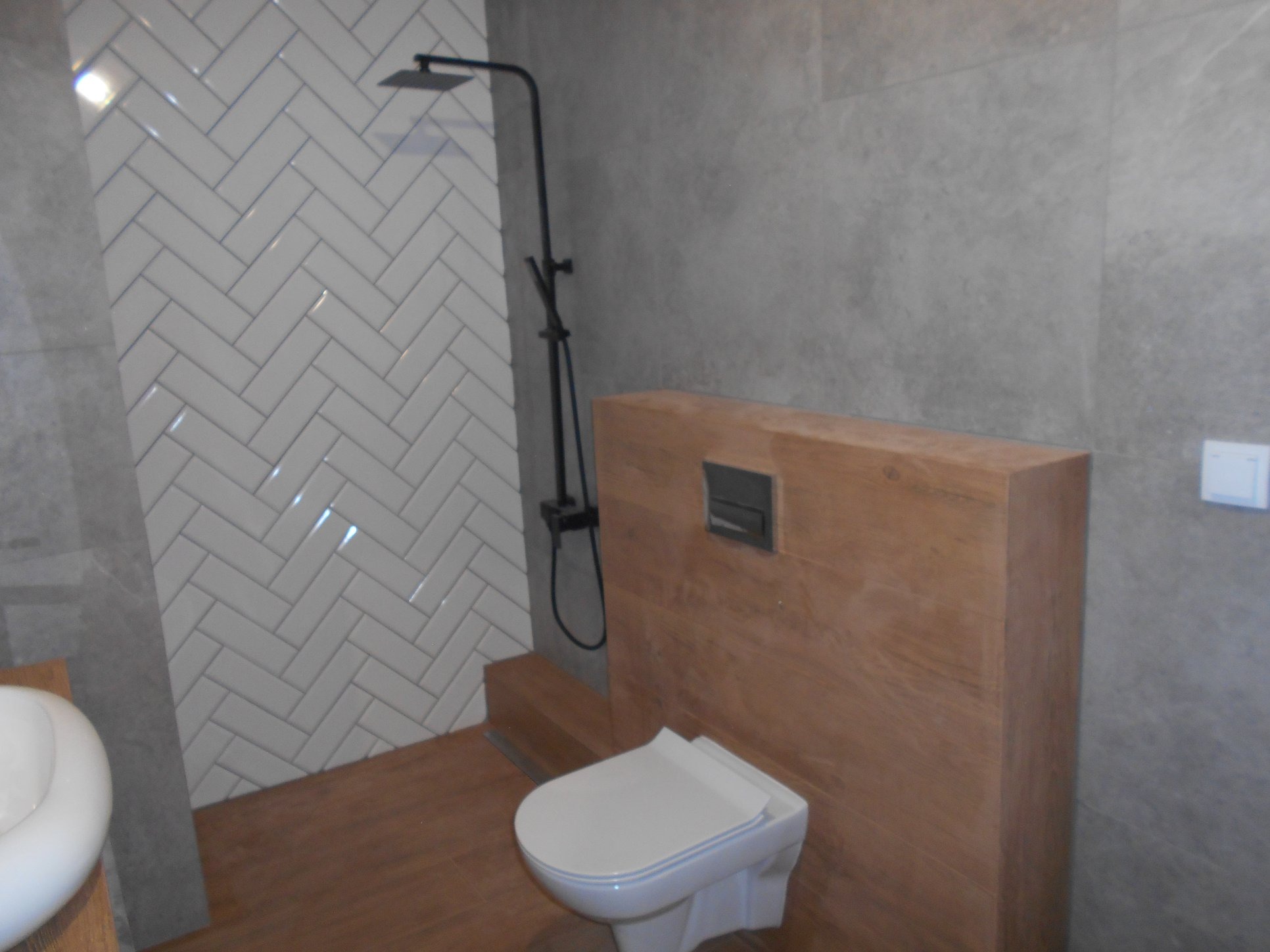 Nowoczesna łazienka z białymi płytkami ułożonymi w jodełkę pod prysznicem, drewnopodobną zabudową toalety oraz deszczownicą w czarnym macie.