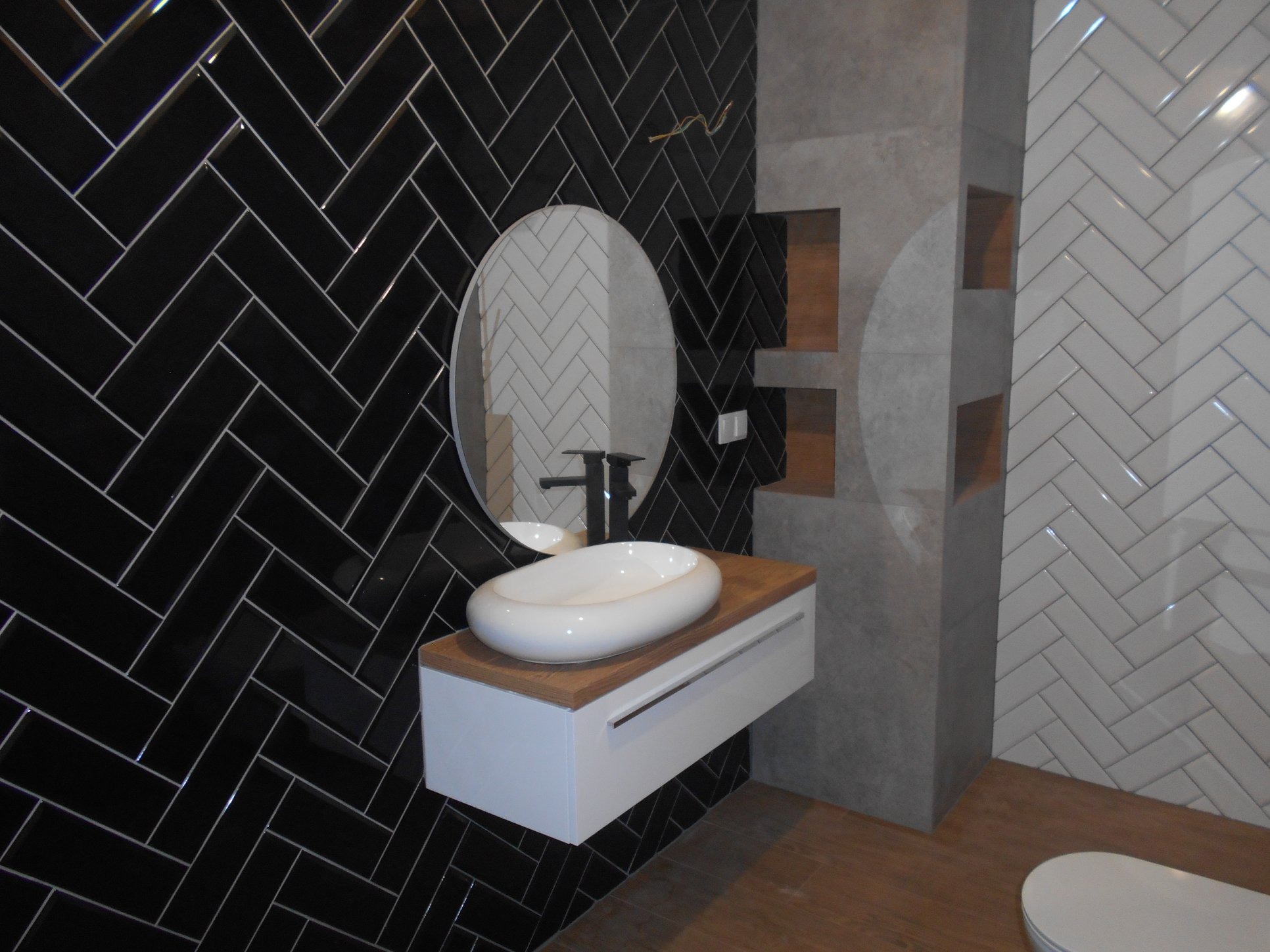Nowoczesna łazienka z czarnymi i białymi płytkami ułożonymi w jodełkę, owalnym lustrem nad białą szafką z drewnianym blatem i umywalką w kształcie misy.