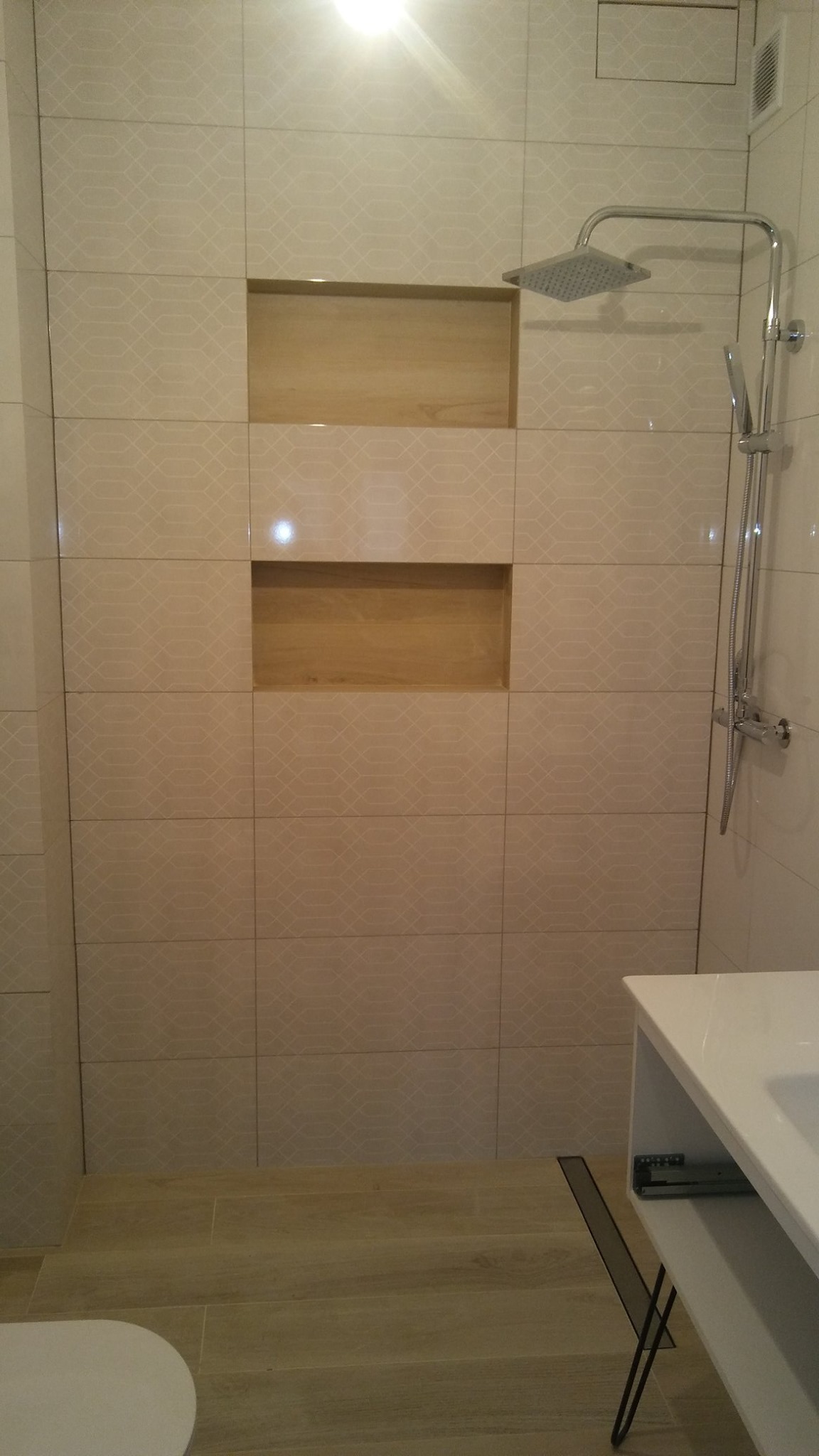 Wykończona łazienka z beżowymi płytkami z delikatnym wzorem, wbudowanymi półkami wyłożonymi drewnem i nowoczesną deszczownicą.