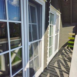 Adam Turowski OKNARO - Białe okna z moskitierami w drewnianym domu, widok z zewnątrz. Refleksy nieba w szybach, drewniana podłoga balkonu. Ujęcie z dołu, jasne światło.