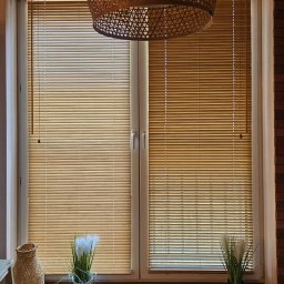 Adam Turowski OKNARO - Beżowe żaluzje poziome zamontowane w oknie PCV, z dekoracjami na parapecie i lampą z wikliny. Widok na okno z bliska, z elementami dekoracyjnymi.