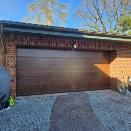 Adam Turowski OKNARO - Brązowa segmentowa brama garażowa z poziomymi panelami, zamontowana w ceglanym garażu z kostką brukową na podjeździe. Widoczne drzewa w tle.