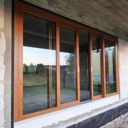 Adam Turowski OKNARO - Duże, przesuwne okno z drewnianą ramą w surowym stanie budynku. Widoczne detale montażu i otoczenie w trakcie budowy. Okno częściowo otwarte.