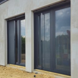 Adam Turowski OKNARO - Dwa okna z zamontowanymi przesuwnymi moskitierami w kolorze grafitowym, widoczne na tle surowej elewacji budynku w trakcie wykończenia.