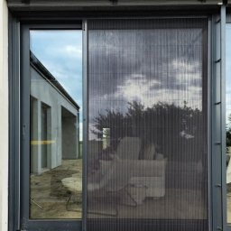 Adam Turowski OKNARO - Szara moskitiera plisowana na oknie przesuwnym, widok z bliska. W tle budynek w stanie surowym i odbicie nieba w szybie. Detal montażu moskitiery.