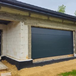 Adam Turowski OKNARO - Nowoczesna, grafitowa brama garażowa segmentowa i drzwi wejściowe w surowym stanie budynku z białego bloczka, widok z zewnątrz, dzień.