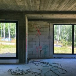 Adam Turowski OKNARO - Surowy stan wnętrza z trzema oknami z czarnymi ramami, widok na zewnątrz na zieleń. Na ścianie oznaczenia instalacji elektrycznej. Kable na podłodze.