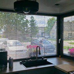 Adam Turowski OKNARO - Kuchnia z oknem narożnym i żaluzjami poziomymi w kolorze ciemnego drewna. Widok na zewnątrz przesłonięty przez żaluzje. Nowoczesny design, czarne ramy okienne.