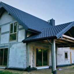 Adam Turowski OKNARO - Surowy dom jednorodzinny z szarą dachówką, widoczne okna z drewnianą ramą i ściany z białego bloczka. Budynek w trakcie budowy, z elementami konstrukcji zadaszenia.