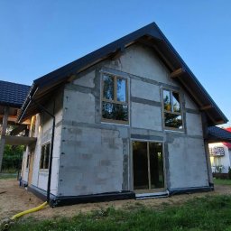 Adam Turowski OKNARO - Surowy dom jednorodzinny w trakcie budowy, z widocznymi oknami i drzwiami w drewnianej ramie, ściany z betonu komórkowego, czarny dach, błękitne niebo w tle.
