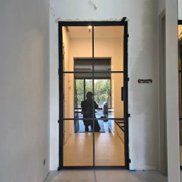 Adam Turowski OKNARO - Nowoczesne, czarne drzwi wewnętrzne z przeszkleniami, widok z korytarza. W tle sylwetka osoby pracującej w pomieszczeniu. Minimalistyczny design, surowy stan wykończenia ścian.