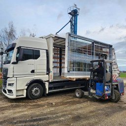 Adam Turowski OKNARO - Transport dużych, przeszklonych elementów konstrukcyjnych na palecie, za pomocą wózka widłowego, do ciężarówki MAN na placu budowy. Widoczna wieża elewatora w tle.