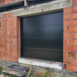 Adam Turowski OKNARO - Czarna brama garażowa segmentowa w surowym otworze muru z cegły. Widoczne detale montażu i izolacji. Ujęcie z bliska, podkreślające solidność konstrukcji.