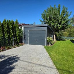 Adam Turowski OKNARO - Nowoczesna, segmentowa brama garażowa w kolorze grafitowym, z poziomymi przetłoczeniami, na tle zadbanego ogrodu i kostki brukowej. Błękitne niebo w tle.