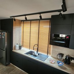 Adam Turowski OKNARO - Nowoczesna kuchnia z drewnianymi żaluzjami poziomymi w oknie nad zlewem. Czarna armatura i oświetlenie szynowe podkreślają minimalistyczny design.