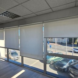 Adam Turowski OKNARO - Białe rolety zwijane zamontowane w oknach biura, widok na ulicę z samochodami. Rolety częściowo opuszczone, wpadające światło dzienne.